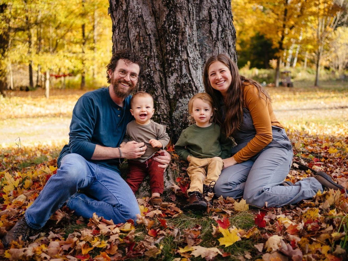 Family portrait in autumn setting, featuring a smiling father holding a baby and a mother sitting beside a toddler. They are surrounded by colorful fall leaves and a large tree, capturing a joyful moment outdoors.