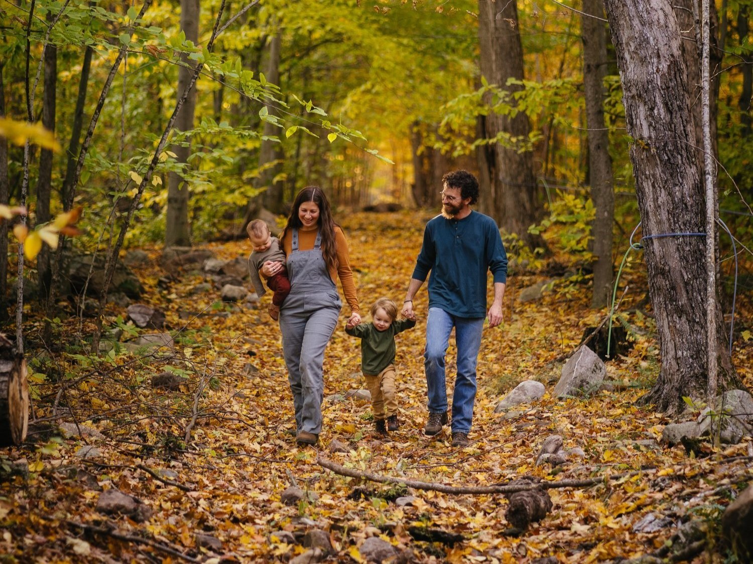 Family enjoying a walk in a colorful autumn forest, surrounded by trees and fallen leaves, with a mother holding a baby and a father walking with a young child.