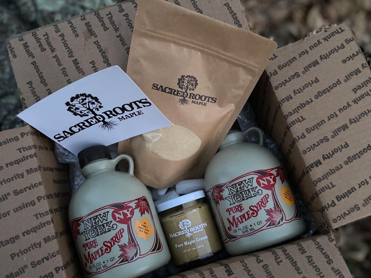 Box of Sacred Roots Maple products featuring two bottles of New York pure maple syrup, a jar of maple cream, and a bag of maple sugar, all presented with branded packaging.